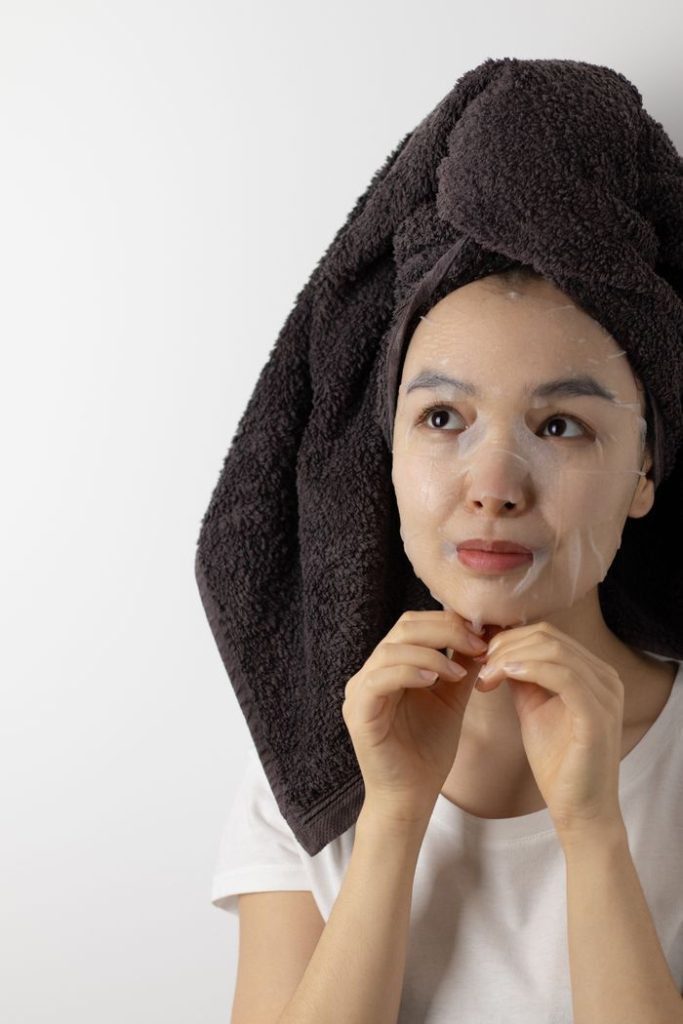 Adult woman with sheet mask and towel on head, enjoying self-care indoors.